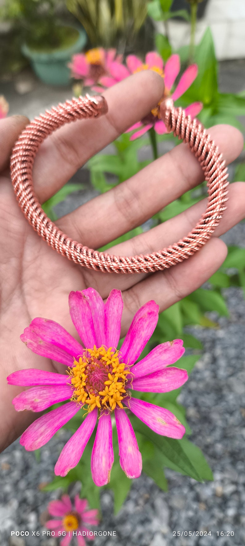 May include: A copper wire bracelet with a braided design. The bracelet is shown on a hand against a background of pink flowers and green foliage.