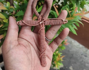 May include: A copper wire Ankh, an ancient Egyptian symbol of life, is held in a hand. The Ankh is made of tightly wound copper wire, giving it a textured appearance.