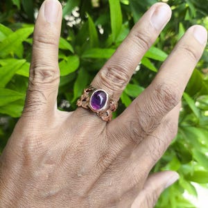 May include: A copper-toned metal ring featuring an oval-shaped, polished purple gemstone set in the center. The band of the ring displays a textured, organic design with intertwined metal elements that wrap around the finger.