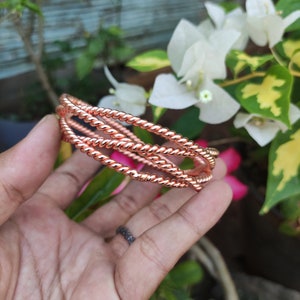 Op de afbeelding: Koperkleurige, gedraaide metalen armbanden. De armbanden zijn gestapeld en lijken te zijn gemaakt van een reeks gedraaide draden. De achtergrond omvat groen gebladerte en witte bloemen.