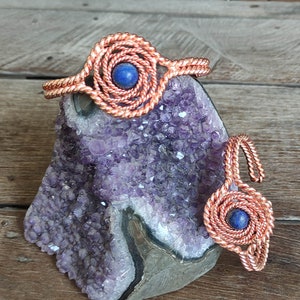 Puede incluir: Un conjunto de pulsera y anillo de color cobre, cada uno con un diseño en espiral y una piedra azul central. La joyería descansa sobre una geoda de amatista púrpura, exhibida sobre una superficie de madera.