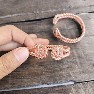Op de afbeelding: Twee koperkleurige armbanden met een gedraaid touwontwerp. Elke armband heeft een open structuur met cirkelvormige, spiraalvormig gewikkelde draaduiteinden. Het metalen oppervlak heeft een warme, roodbruine tint en een textuur.