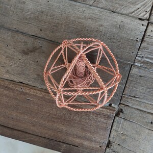 May include: A copper-colored geometric sculpture with a spherical shape. The sculpture is made of twisted wire, creating an open, cage-like design. The sculpture sits on a weathered wooden surface.