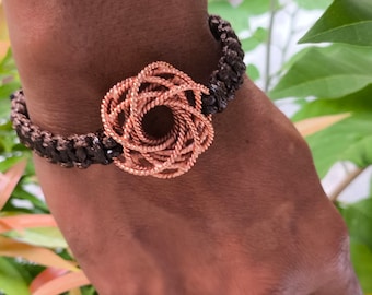 May include: A copper wire bracelet with a woven flower design. The bracelet is on a person's wrist and the background is blurred.