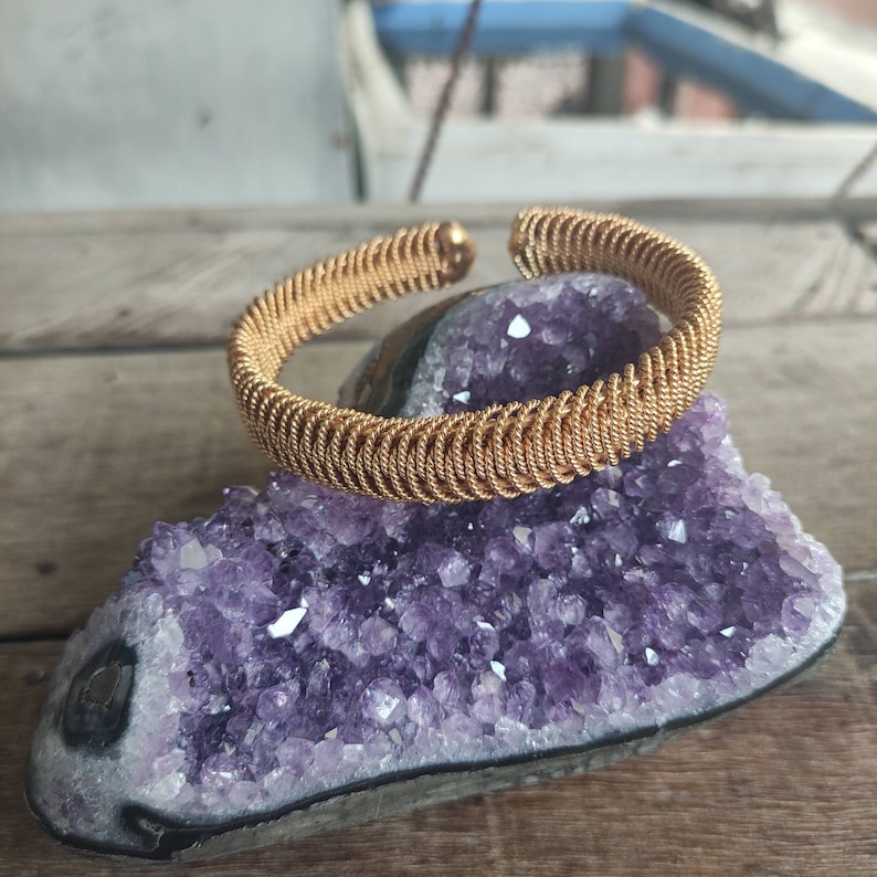 May include: A gold-toned wire bangle bracelet, resting on a bed of purple amethyst crystals.
