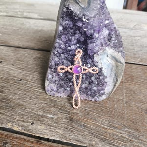 May include: A cross pendant crafted from twisted copper wire featuring a single round purple gemstone set at the center. The cross is positioned against a large, jagged amethyst geode with deep purple crystalline structures throughout the stone.