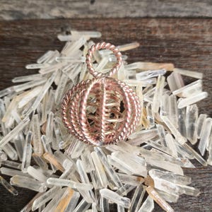Peut inclure: Un pendentif sphérique de couleur cuivrée avec un motif en spirale, suspendu au-dessus d'un lit de cristaux de quartz clairs. Le pendentif a une boucle en haut pour l'accrocher. Les cristaux sont de tailles et de formes variées, reposant sur une surface en bois.