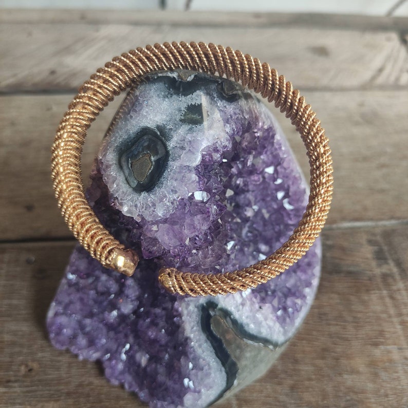 May include: A gold-toned metal bangle bracelet with a twisted wire design. The bracelet is displayed on a purple amethyst geode.