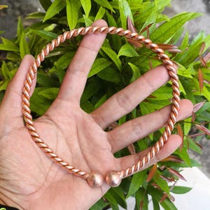 May include: A copper-colored, twisted-wire necklace with two rounded end pieces. The necklace is held in a hand against a backdrop of green foliage. The necklace appears to be approximately 6 inches in diameter.