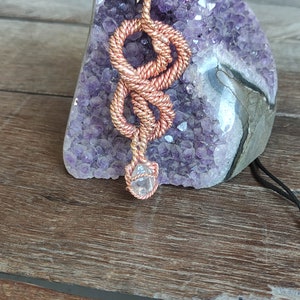 May include: A copper wire wrapped pendant with a clear crystal at the bottom. The pendant is hanging from a black cord and is displayed on a purple amethyst geode.