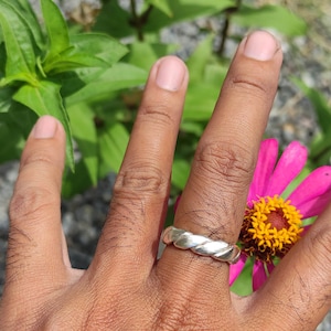 May include: A silver ring with a twisted design, worn on a person's finger. The ring is in focus, while the background is blurred and shows green leaves and a pink flower.