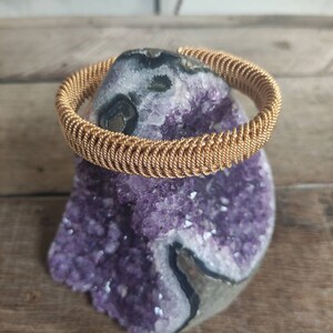 May include: A gold-colored wire bracelet with a braided design. The bracelet is displayed on a purple amethyst geode.