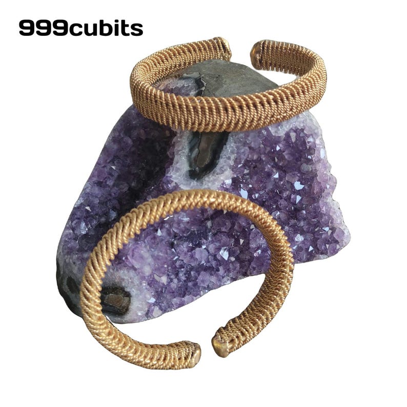 May include: Two gold wire bracelets on a purple amethyst crystal cluster. The bracelets are made of twisted wire and are both open ended.