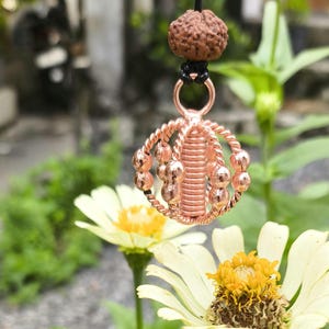 Puede incluir: Un colgante de cobre con un diseño en espiral y pequeñas cuentas. El colgante está colgado de un cordón negro con una cuenta marrón en la parte superior.