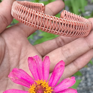 May include: A copper wire bracelet with a woven design. The bracelet is shown on a hand against a background of pink flowers.
