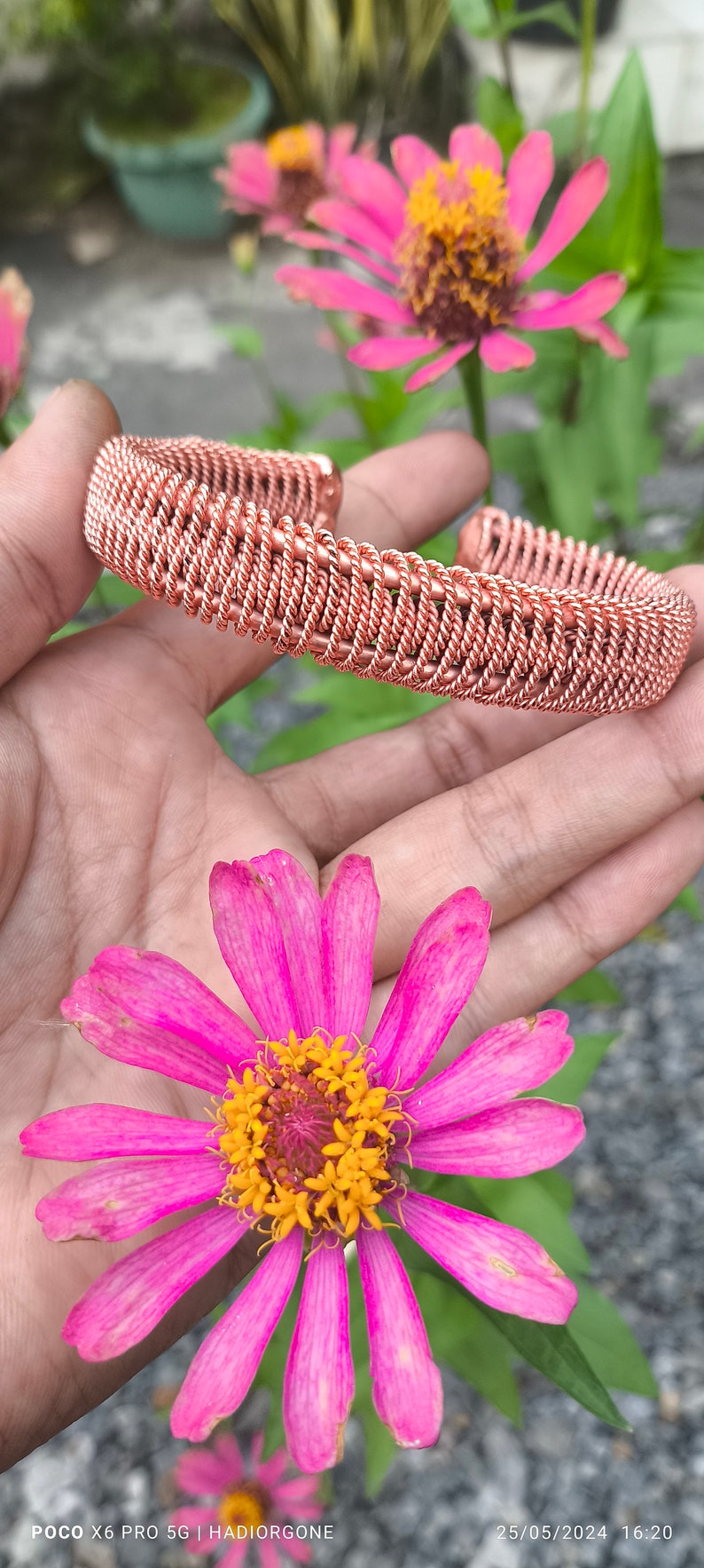 May include: A copper wire bracelet with a woven design. The bracelet is shown on a hand against a background of pink flowers.