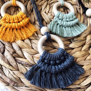 May include: Three wooden ring toys with macrame fringe. The rings are natural wood and the fringe is yellow, green, and gray.