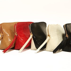 May include: A collection of five rectangular pouches with a crocodile-embossed texture, each featuring a gold-tone zipper closure and a matching pull tab. The pouches are arranged in a row and come in tan, red, black, white, and black colors.