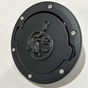 May include: Black round gas cap with a detailed Medusa head design. The cap has a hinged opening and is secured with silver screws.