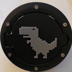 Può includere: Coperchio del bocchettone del carburante nero e grigio con un design a dinosauro pixelato. Il coperchio è rotondo con un bordo nero ed è fissato con viti argentate. Il dinosauro è grigio e centrato sullo sfondo nero.