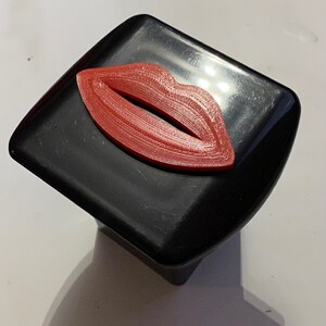 Puede incluir: Una caja cuadrada negra con un diseño de labios rojo impreso en 3D en la parte superior.