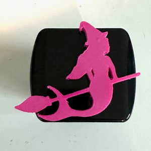 May include: A pink witch silhouette on a black background. The witch is riding a broomstick and has a pointed hat.