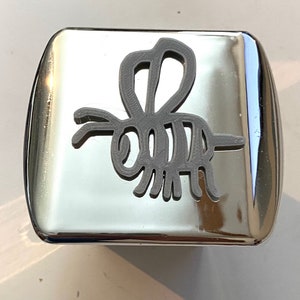 Puede incluir: Una tapa de enganche cuadrada plateada con un diseño de abeja impreso en 3D gris.