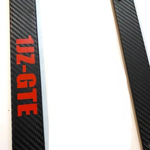 Pode incluir: Moldura de placa de matrícula em fibra de carbono preta com um logotipo "1JZ-GTE" vermelho.