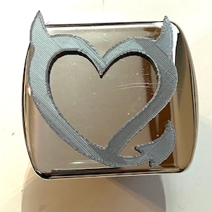 Puede incluir: Una tapa de enganche de metal plateado con un diseño impreso en 3D gris de un corazón con cuernos de diablo.