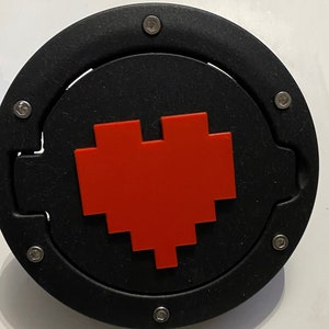Può includere: Un tappo di benzina rotondo nero con un design a cuore pixelato rosso. Il tappo ha diverse viti argentate attorno al bordo.