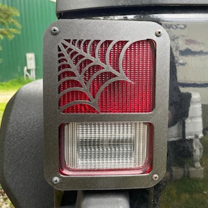 May include: Black metal Jeep tail light cover with a spider web cut-out design. The cover is installed on a black Jeep.