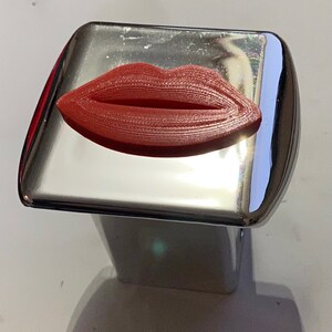Puede incluir: Un objeto rojo con forma de labios, impreso en 3D, descansa sobre una superficie plateada brillante. Los labios tienen líneas horizontales detalladas, dándoles una apariencia texturizada. La superficie refleja la luz.