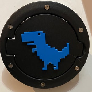 Può includere: Un tappo serbatoio rotondo nero con un dinosauro pixelato blu. Il tappo serbatoio ha sei viti argentate attorno al bordo.