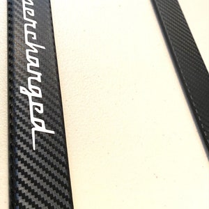 May include: Black carbon fiber license plate frame with the word "Supercharged" in white lettering.