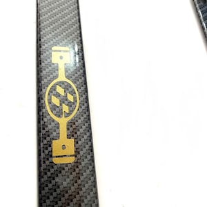 May include: A black carbon fiber license plate frame with a gold logo of a piston and a stylized gear.