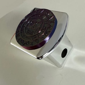 Puede incluir: Una tapa de enganche cromada con un diseño de calavera y huesos cruzados en negro y morado. El diseño presenta un círculo con números alrededor del exterior y una calavera en el centro.