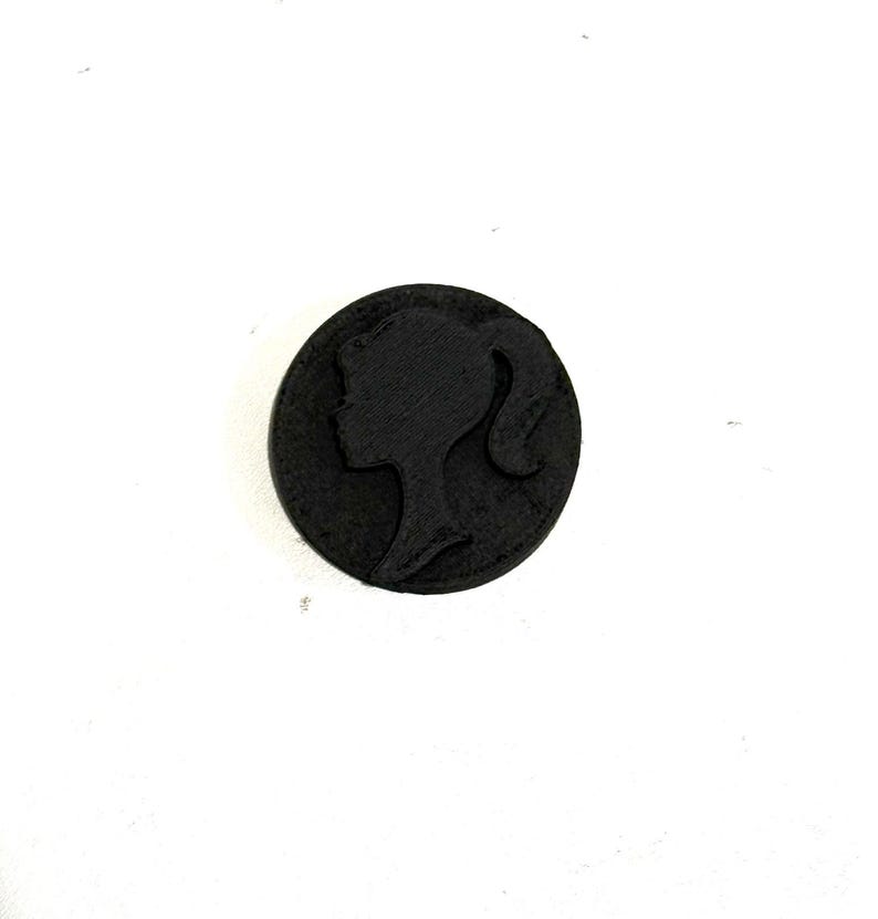 May include: A black circular button with a raised silhouette of a woman's face.