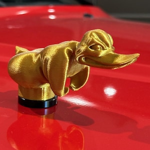 Könnte beinhalten: Ein goldfarbener Enten-Kappen-Schmuck für ein Auto. Die Ente hat eine strukturierte Oberfläche und befindet sich auf einer roten Motorhaube.