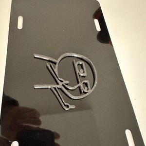 May include: A black metal license plate with a silver 3D design of a face with a surprised expression.