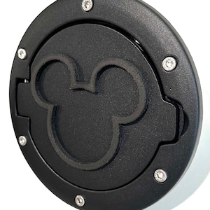 Puede incluir: Una tapa de puerta de combustible negra y circular con un contorno en relieve de la cabeza de Mickey Mouse. La tapa está asegurada con tornillos plateados y tiene un diseño con bisagras. El producto es de metal.