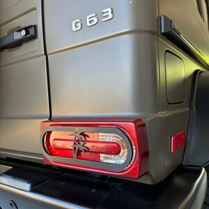 Könnte beinhalten: Nahaufnahme des Hecklichts eines dunkelgrünen SUVs. Das Rücklicht ist rot und hat einen schwarzen Palmen-Ausschnitt. Das Fahrzeug hat ein silbernes "G 63" Emblem am Heck.