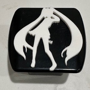 May include: A black and white car emblem featuring a silhouette of a female anime character with long hair. The character is standing with one leg bent and the other straight, with her arms raised above her head.