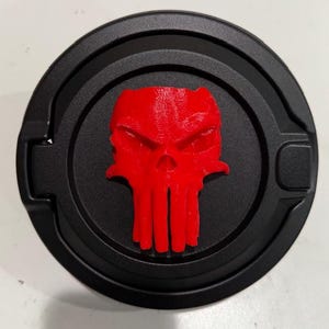 May include: A black, circular object with a raised, red skull emblem in the center. The skull has a menacing expression. The object appears to be a decorative car accessory.