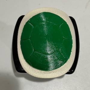 Könnte beinhalten: Ein 3D-gedrucktes Objekt in Form eines Schildkrötenpanzers. Die Schale ist grün mit einer strukturierten Oberfläche, umgeben von einem cremefarbenen Rand. Der Sockel ist schwarz und gebogen, was dem Objekt ein abgerundetes Aussehen verleiht. Das Design ist detailliert und geometrisch.