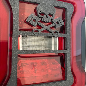 May include: Black skull and crossbones design with pistons on a red car taillight cover. The cover is made of a textured black material.