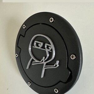 May include: A black gas cap with a silver 3D printed design of a stick figure giving the middle finger.