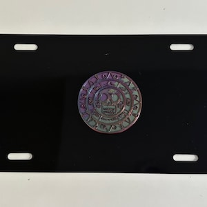 Puede incluir: Una placa de matrícula negra con cuatro orificios de montaje. Una moneda circular e iridiscente con un diseño de calavera está centrada en la placa. La moneda presenta tonos morados, verdes y azules.