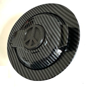 May include: A black carbon fiber gas cap with a peace sign emblem. The cap has a textured surface and a raised lip around the edge.