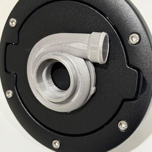 May include: A black, circular fuel door cover with a silver-colored turbocharger design. The cover is secured with six silver screws. The turbocharger is a detailed, 3D-printed design.