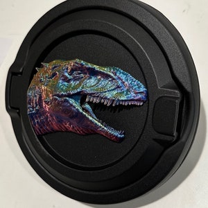 May include: A black round gas cap with a 3D iridescent dinosaur head in the center. The dinosaur is facing right and has a wide open mouth with sharp teeth.